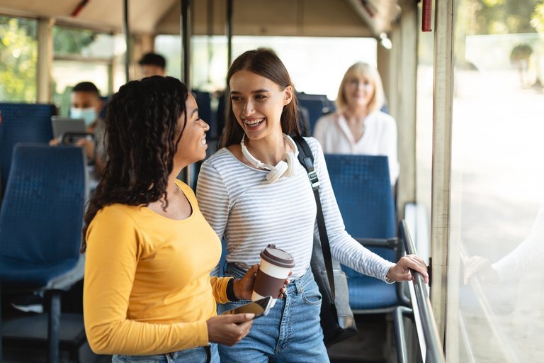 Public Transportaton. Two smiling young women standing at autobus, talking and looking at each other, holding takeaway coffee cup and cell phone, students enjoying ride or travel together in vehicle