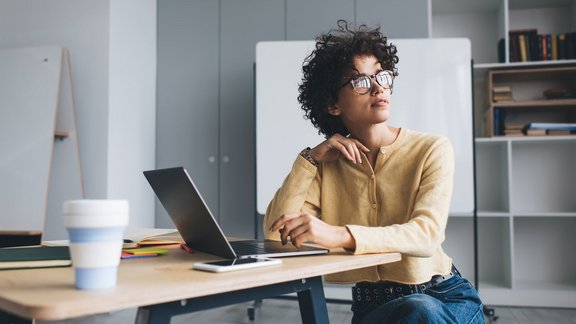 Bewerbungs- und PC Coaching Millennial Geschäftsfrau arbeitet nachdenklich an einem Schreibtisch mit geöffnetem Laptop arbeitet. Sie trägt Brille und sitzt in einem Open-Space-Büro.