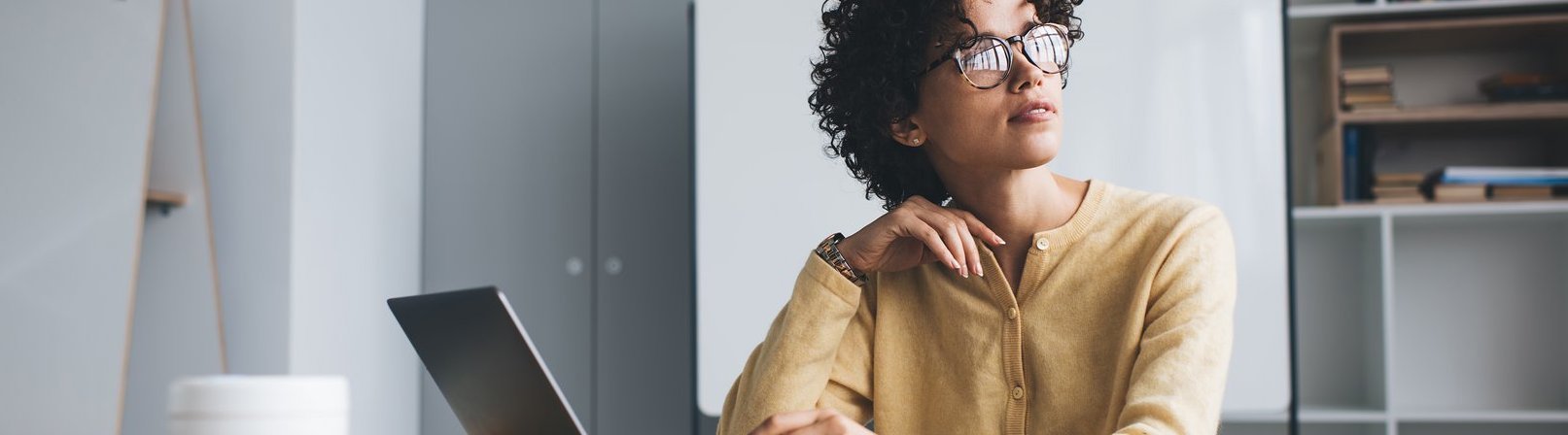 Bewerbungs- und PC Coaching inkl. Jobcenter digital Millennial Geschäftsfrau arbeitet nachdenklich an einem Schreibtisch mit geöffnetem Laptop arbeitet. Sie trägt Brille und sitzt in einem Open-Space-Büro.