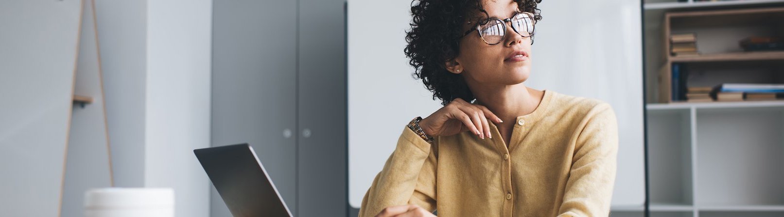 Gewusst wie – sicherer Umgang mit digitalen Medien Millennial Geschäftsfrau arbeitet nachdenklich an einem Schreibtisch mit geöffnetem Laptop arbeitet. Sie trägt Brille und sitzt in einem Open-Space-Büro.