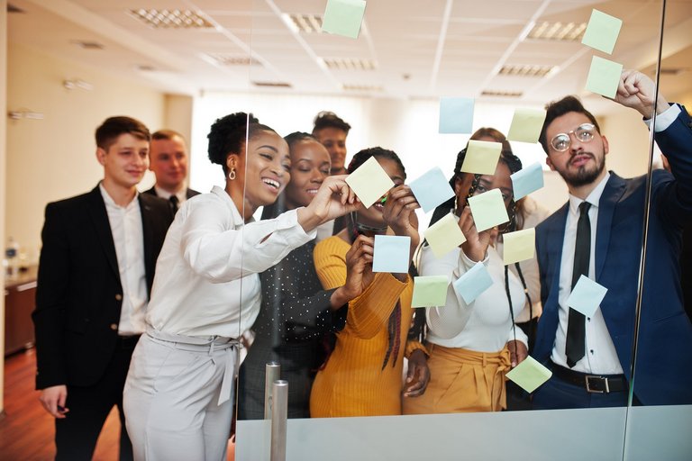 Großes Team aus Männern und Frauen steht in einer Business-Situation vor einer Glaswand bedeckt mit Post-its.