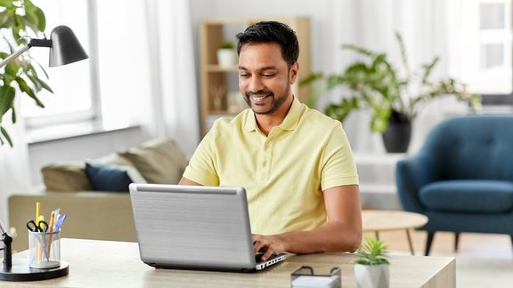 Ein glücklicher, lächelnder indischer Mann, der mit einem Laptop im Homeoffice arbeitet. Er trägt ein gelbes Hemd.