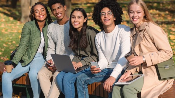 Fröhliche internationale Teenager-Freunde sitzen auf einer Bank mit ihren Geräten und verbringen Zeit zusammen im Park.